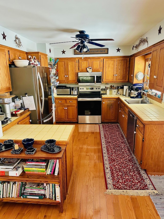 kitchen featuring light wood finished floors, brown cabinetry, tile countertops, stainless steel appliances, and a sink