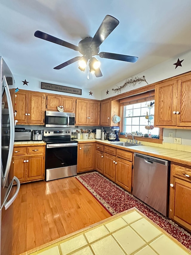 kitchen with a sink, a ceiling fan, appliances with stainless steel finishes, tile counters, and brown cabinetry