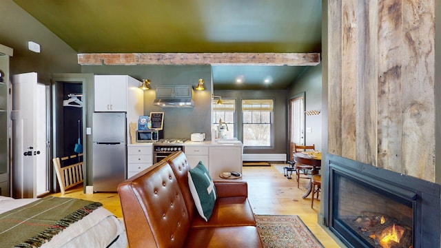 kitchen featuring vaulted ceiling with beams, light wood-style flooring, appliances with stainless steel finishes, a glass covered fireplace, and white cabinetry