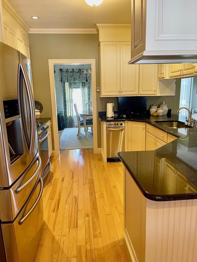 kitchen with light wood finished floors, appliances with stainless steel finishes, dark stone countertops, crown molding, and a sink