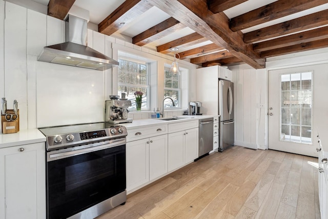 kitchen with a sink, appliances with stainless steel finishes, wall chimney range hood, light wood-type flooring, and beamed ceiling