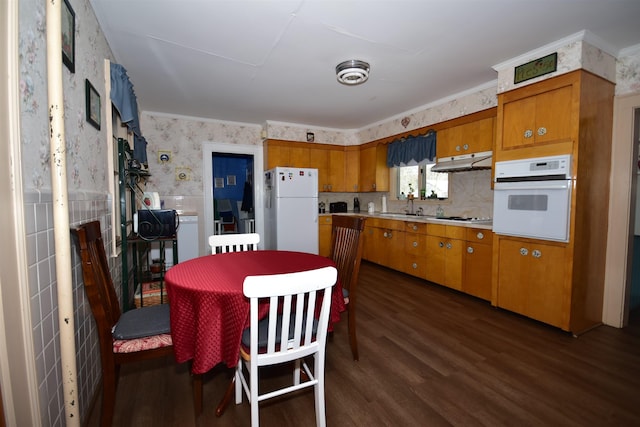 kitchen with white appliances, wallpapered walls, dark wood-style floors, brown cabinets, and under cabinet range hood