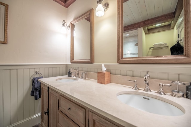 full bath with double vanity, beam ceiling, a sink, and wainscoting