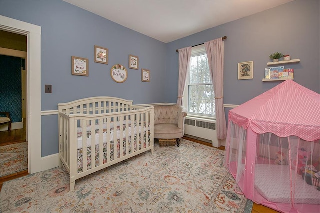 bedroom with a nursery area, radiator heating unit, wood finished floors, and baseboards