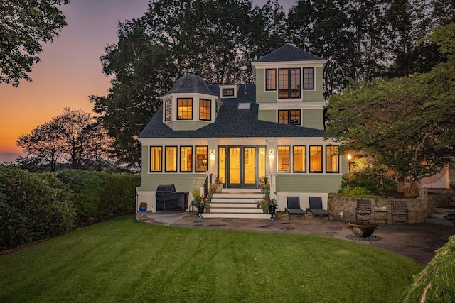 rear view of property featuring entry steps, a shingled roof, a lawn, an attached garage, and a patio area