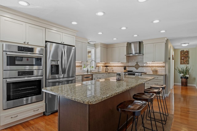 kitchen featuring stainless steel appliances, tasteful backsplash, light wood-style flooring, wall chimney range hood, and a kitchen bar