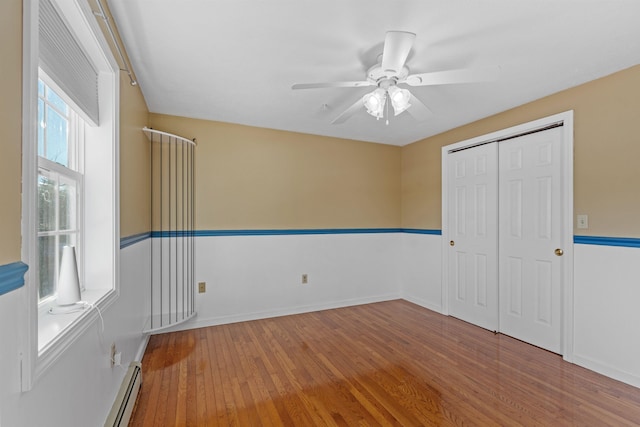 unfurnished bedroom featuring a baseboard heating unit, wood finished floors, a ceiling fan, baseboards, and a closet