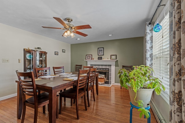 dining room with baseboards, a ceiling fan, a tile fireplace, baseboard heating, and light wood-style floors