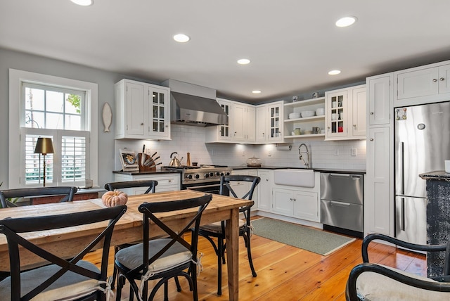 kitchen with a sink, appliances with stainless steel finishes, light wood-type flooring, wall chimney exhaust hood, and dark countertops