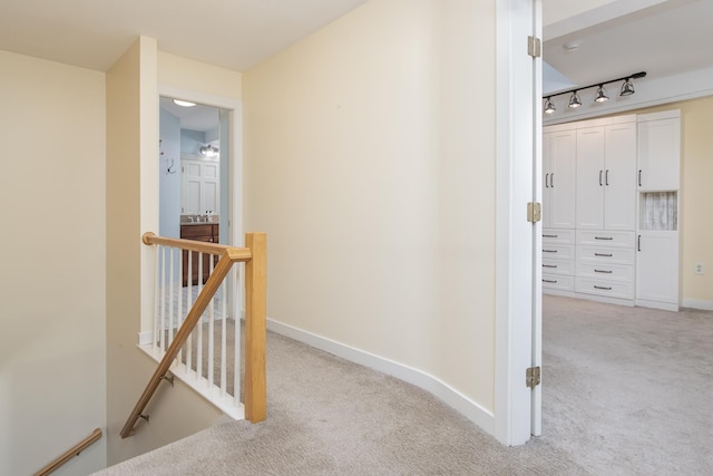 hall featuring baseboards, rail lighting, an upstairs landing, and light colored carpet