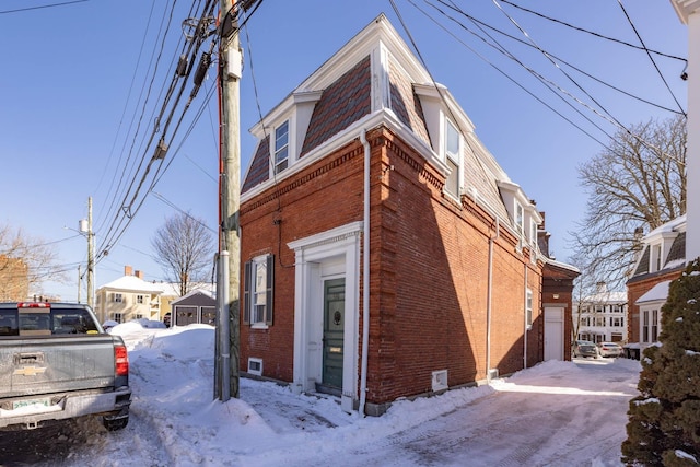 view of snow covered exterior with a garage, brick siding, and mansard roof