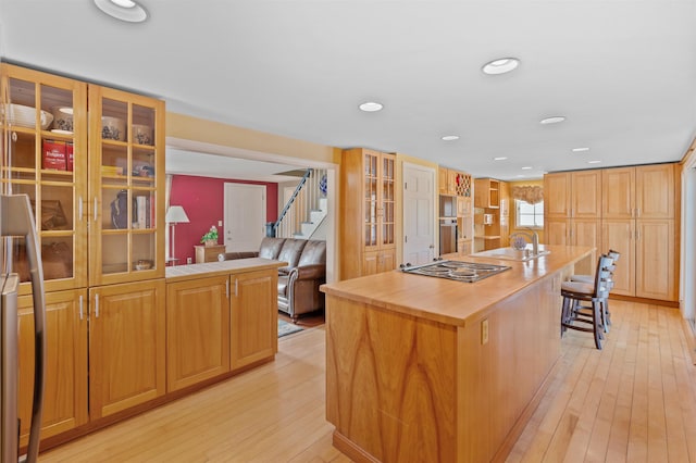 kitchen with electric stovetop, light wood-style flooring, a kitchen breakfast bar, an island with sink, and glass insert cabinets