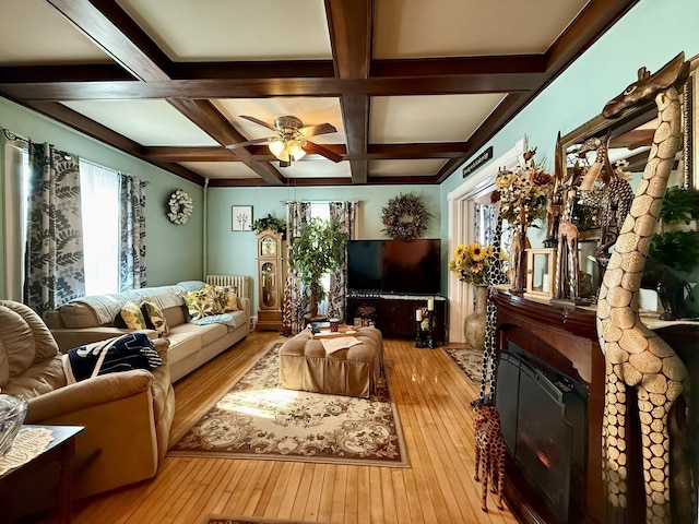 living area with coffered ceiling, a glass covered fireplace, ceiling fan, wood-type flooring, and beam ceiling