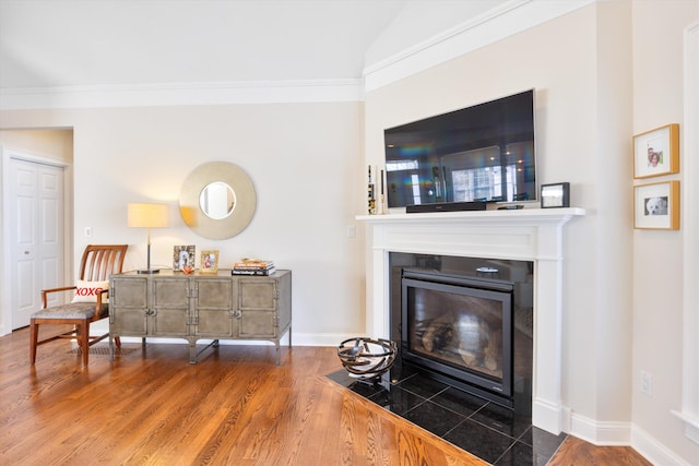 living area featuring a fireplace with flush hearth, wood finished floors, baseboards, vaulted ceiling, and crown molding