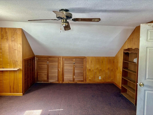 bonus room with wooden walls, lofted ceiling, ceiling fan, dark colored carpet, and a textured ceiling