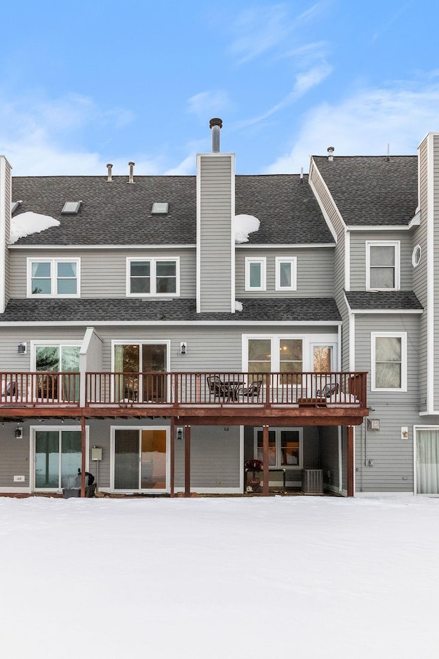 snow covered rear of property with a shingled roof, a chimney, and cooling unit