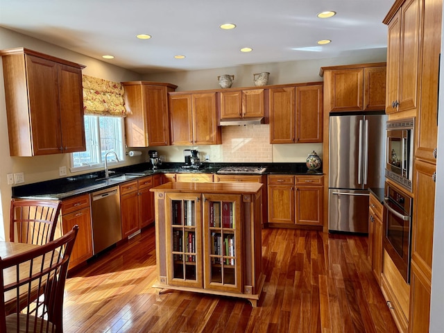 kitchen with brown cabinetry, dark wood-style floors, stainless steel appliances, under cabinet range hood, and a sink