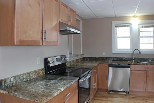 kitchen with dark stone counters, a sink, a paneled ceiling, under cabinet range hood, and appliances with stainless steel finishes