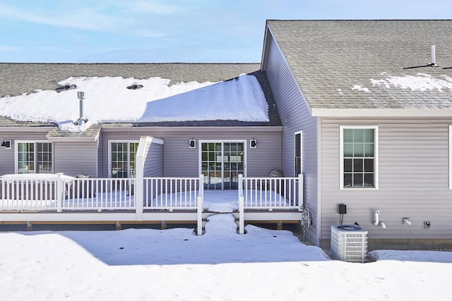 snow covered back of property with central AC unit, a deck, and roof with shingles