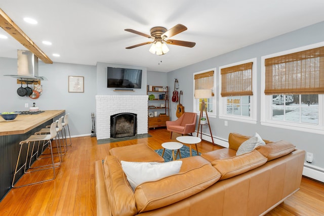 living area with a baseboard heating unit, wood finished floors, a ceiling fan, baseboards, and a brick fireplace