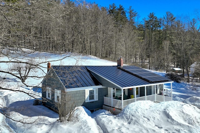 snow covered property with a standing seam roof, metal roof, covered porch, roof mounted solar panels, and a chimney