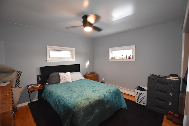 bedroom with a ceiling fan, multiple windows, a baseboard heating unit, and wood finished floors