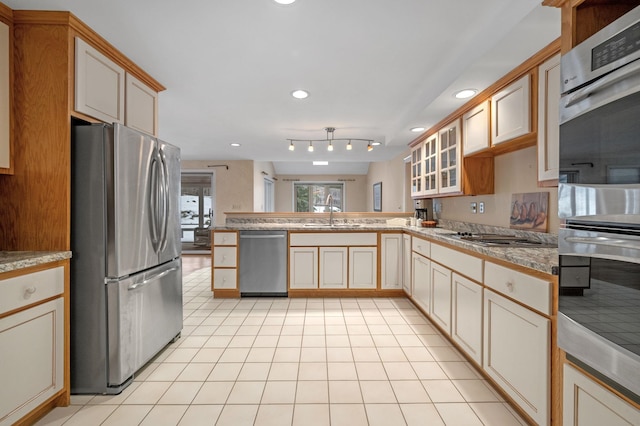 kitchen featuring light tile patterned floors, a peninsula, a sink, appliances with stainless steel finishes, and glass insert cabinets