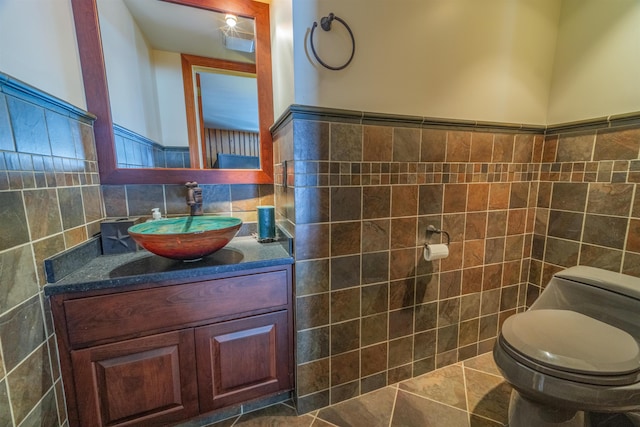 half bath with tile patterned flooring, toilet, a wainscoted wall, vanity, and tile walls