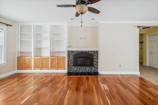 unfurnished living room featuring ceiling fan, wood finished floors, baseboards, ornamental molding, and a tiled fireplace