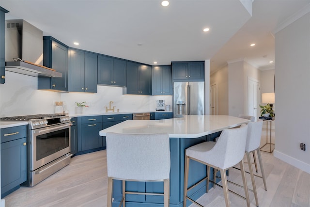 kitchen featuring blue cabinetry, stainless steel appliances, wall chimney range hood, light wood-type flooring, and a kitchen bar