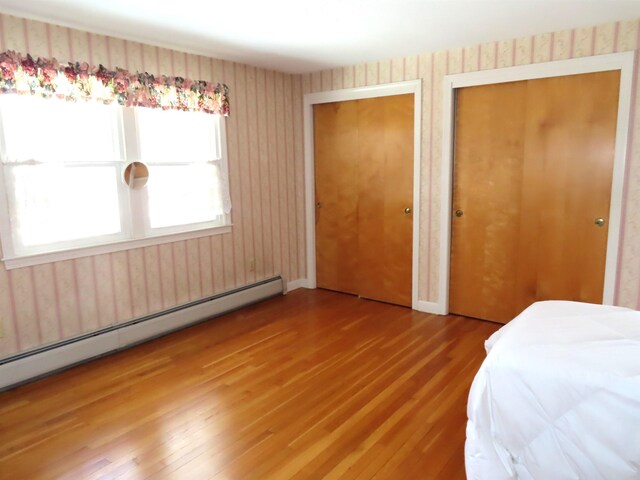 unfurnished bedroom featuring multiple closets, a baseboard radiator, wood finished floors, and wallpapered walls