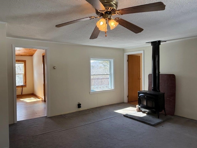 unfurnished living room featuring carpet floors, ornamental molding, a wood stove, ceiling fan, and a textured ceiling