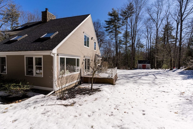 view of snow covered exterior with an outbuilding, a chimney, a storage unit, a shingled roof, and a deck