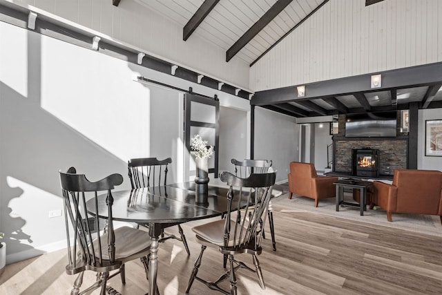 dining area with wood finished floors, high vaulted ceiling, beam ceiling, a stone fireplace, and a barn door