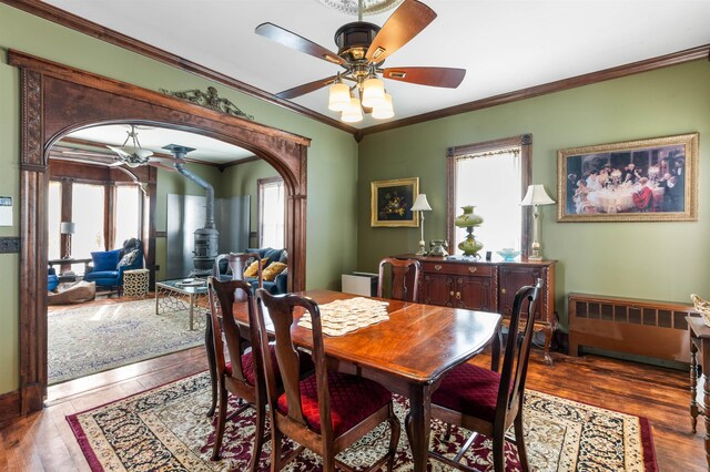 dining space with arched walkways, a ceiling fan, hardwood / wood-style flooring, a wood stove, and crown molding