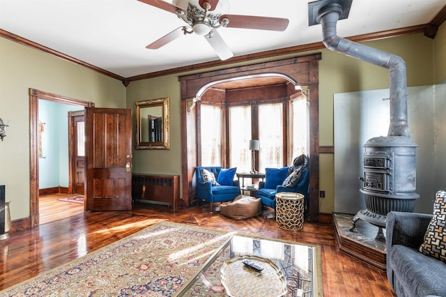 living area featuring baseboards, ceiling fan, ornamental molding, wood finished floors, and a wood stove