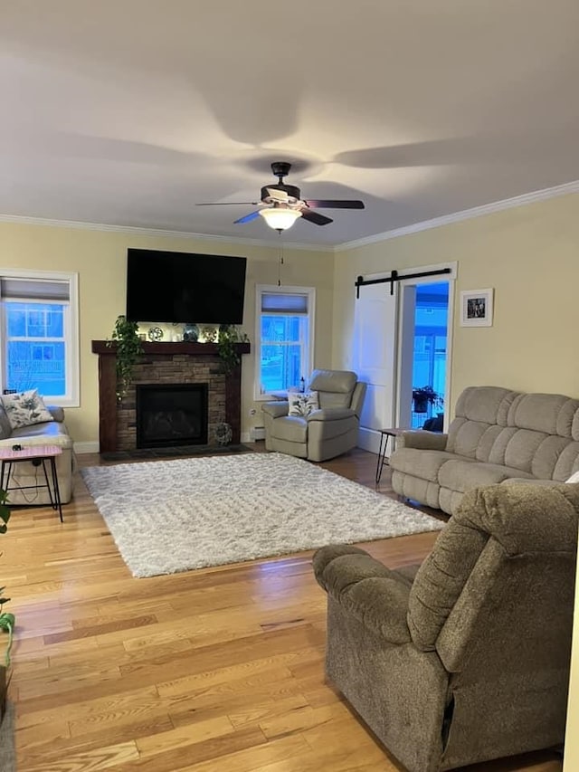 living room with a ceiling fan, a barn door, light wood-style floors, a stone fireplace, and crown molding