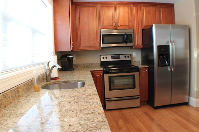 kitchen featuring light wood finished floors, light stone countertops, brown cabinets, appliances with stainless steel finishes, and a sink