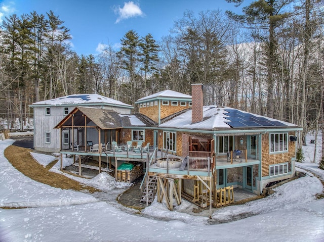 snow covered back of property featuring a wooden deck, stairway, roof mounted solar panels, and a chimney