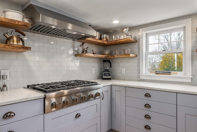 kitchen with wall chimney range hood, light stone countertops, stainless steel gas cooktop, decorative backsplash, and open shelves