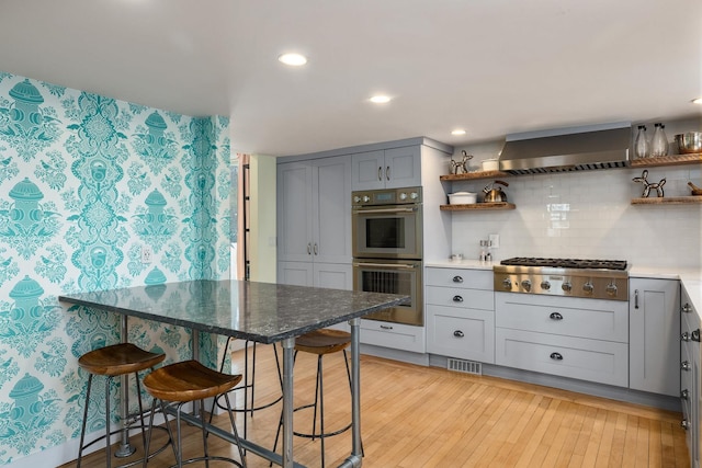 kitchen with gray cabinetry, open shelves, stainless steel appliances, wall chimney exhaust hood, and light wood finished floors