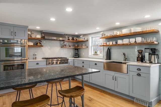 kitchen featuring open shelves, light wood-style floors, appliances with stainless steel finishes, a kitchen bar, and wall chimney exhaust hood