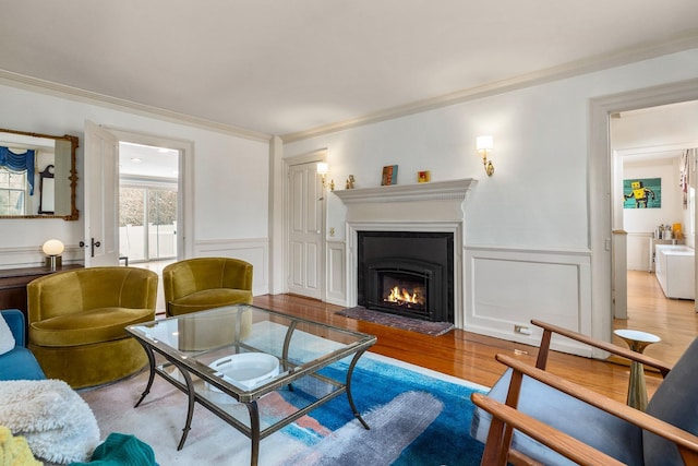 living area with wood finished floors, ornamental molding, a lit fireplace, wainscoting, and a decorative wall