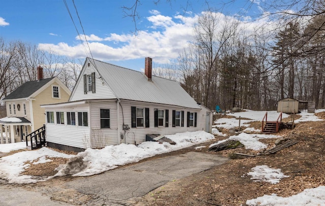 snow covered property featuring an outbuilding, aphalt driveway, a chimney, a storage shed, and metal roof