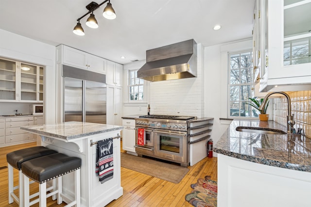 kitchen with wall chimney range hood, dark stone counters, white cabinetry, high end appliances, and a sink