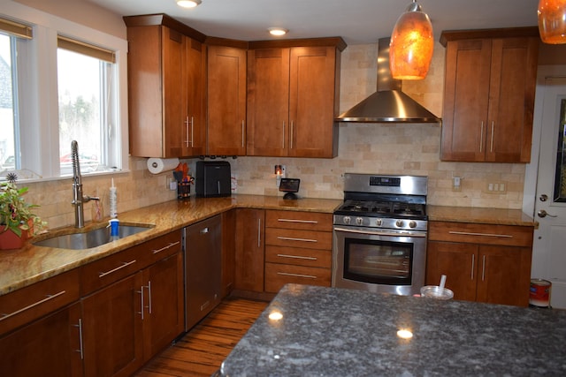 kitchen featuring wall chimney range hood, dark stone counters, appliances with stainless steel finishes, brown cabinetry, and a sink