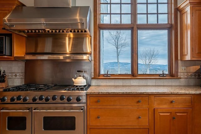 kitchen with decorative backsplash, double oven range, wall chimney exhaust hood, and light stone countertops