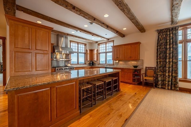 kitchen featuring stone counters, brown cabinets, light wood finished floors, wall chimney exhaust hood, and tasteful backsplash