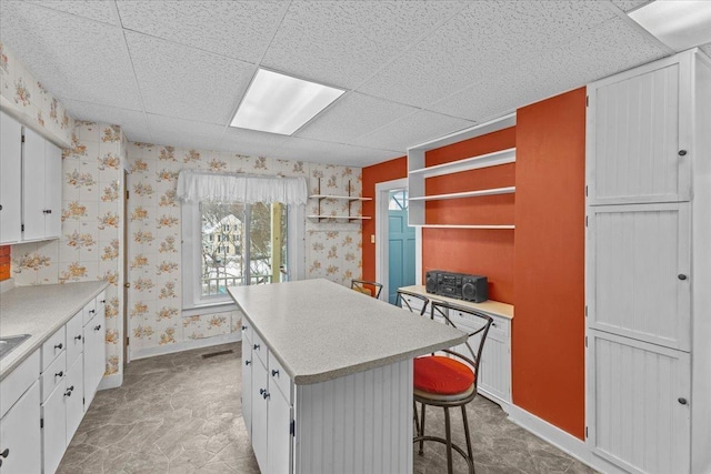 kitchen featuring wallpapered walls, a drop ceiling, white cabinetry, and a kitchen island