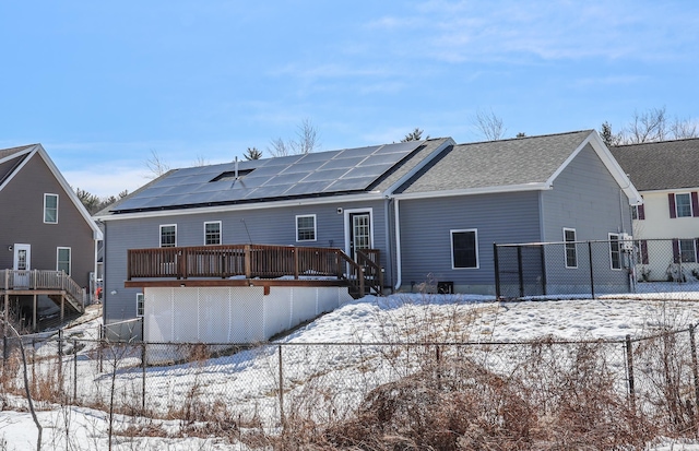 snow covered property with solar panels, a shingled roof, a deck, and fence